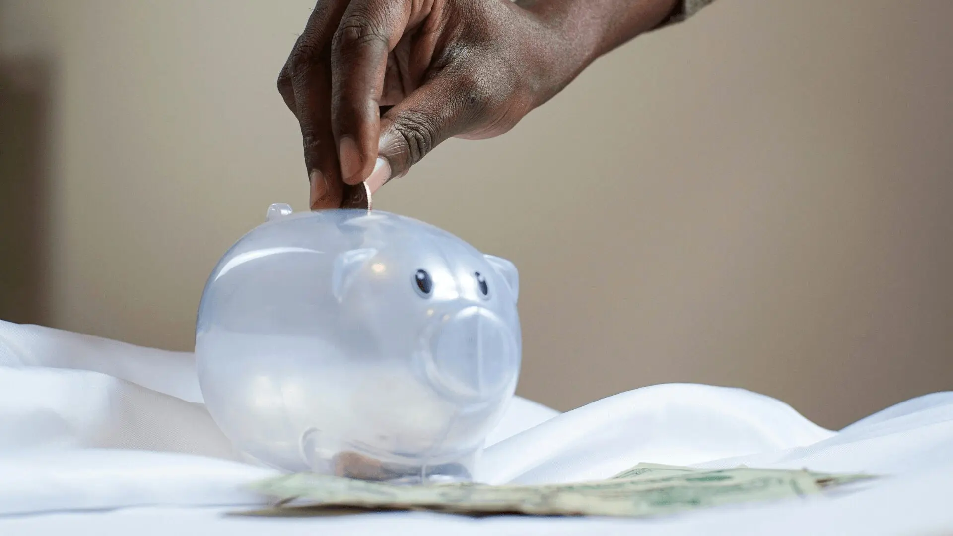 A dark-skinned hand dropping a coin into a translucent piggy bank, with paper money visible underneath.