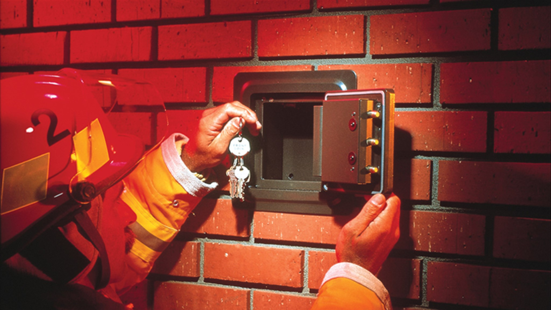 A firefighter in a red helmet and jacket inserting keys into an open KnoxBox mounted in a red brick wall