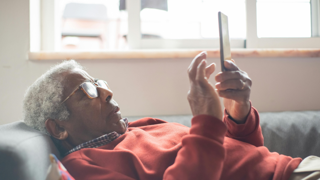 An elderly man with glasses wearing a red sweater comfortably using a smartphone while relaxing on a couch.