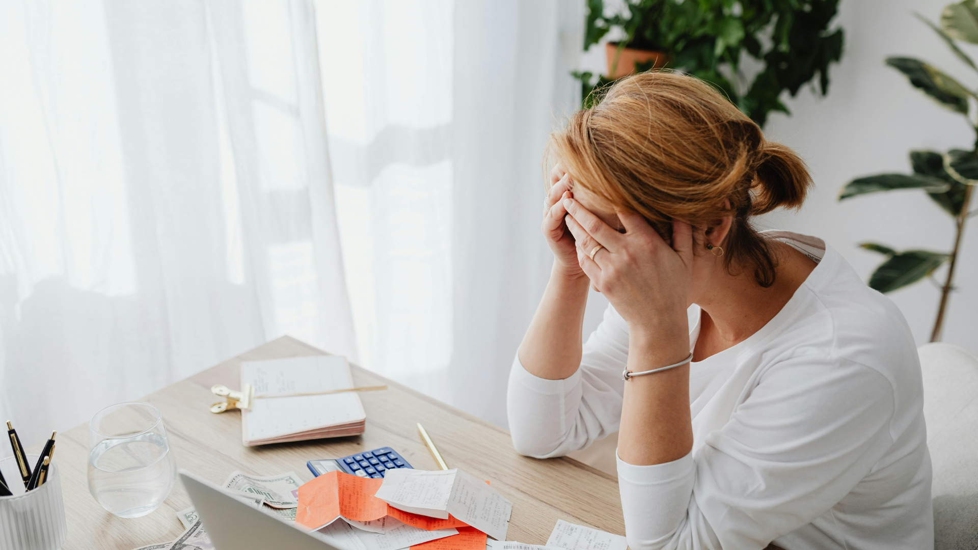 A woman with red hair looking stressed, holding her head in her hands while looking at papers on a desk with a calculator.