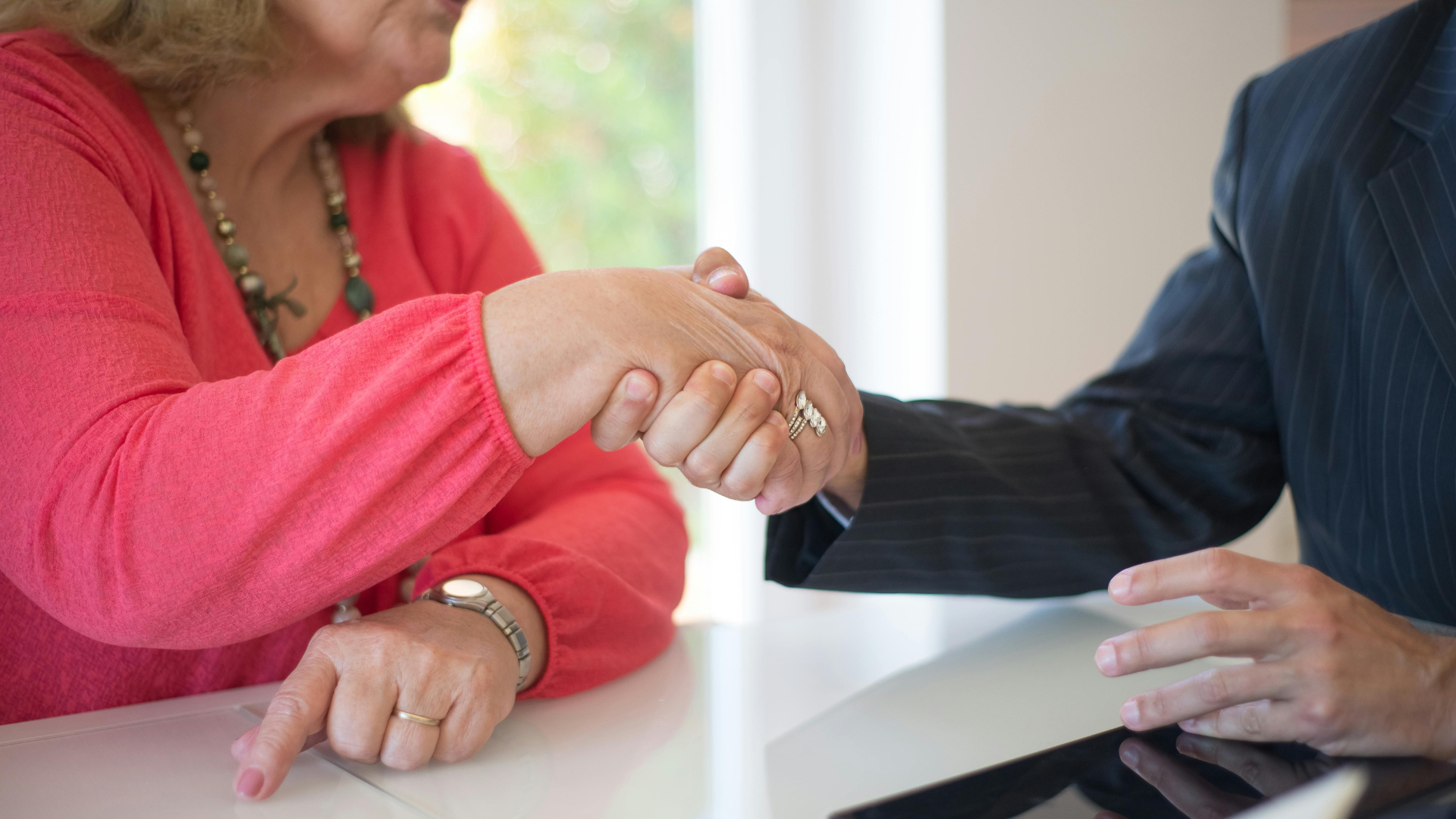 Two people shaking hands across a table, one a senior with rings, representing a financial agreement.