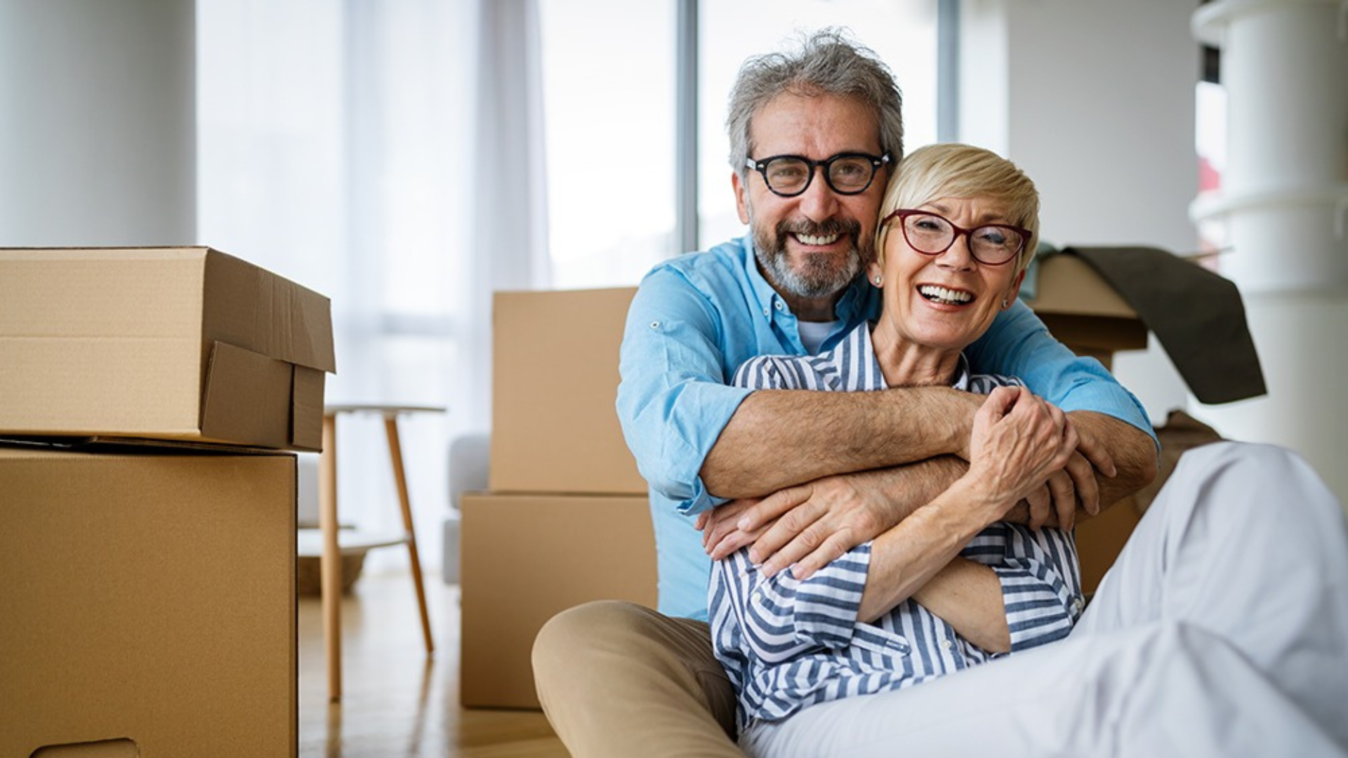A happy senior couple, smiling and embracing, sitting on the floor surrounded by moving boxes.
