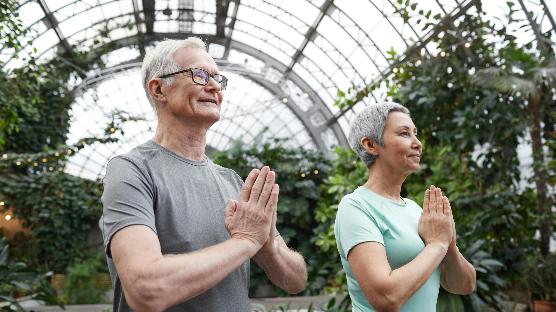 A senior man and woman with hands in prayer position, practicing yoga or meditation in a lush indoor garden setting.