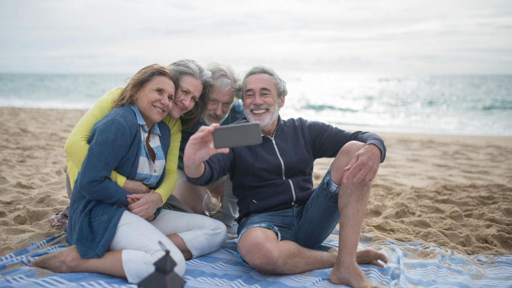 A group of four smiling senior friends taking a selfie together on a beach.