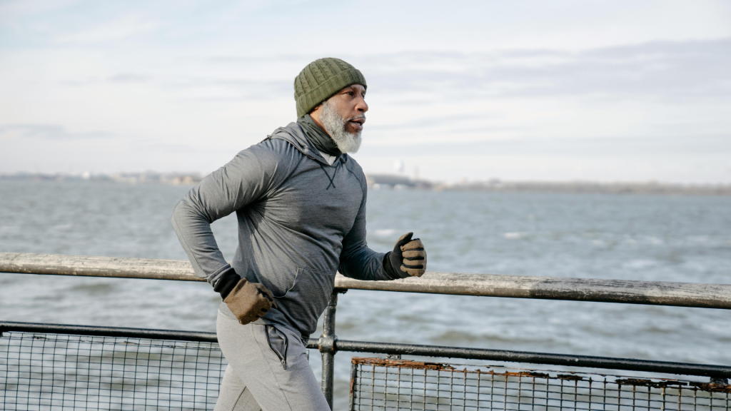 A senior man with a white beard and hat, wearing a gray sweatshirt and gloves, jogging on a waterfront boardwalk