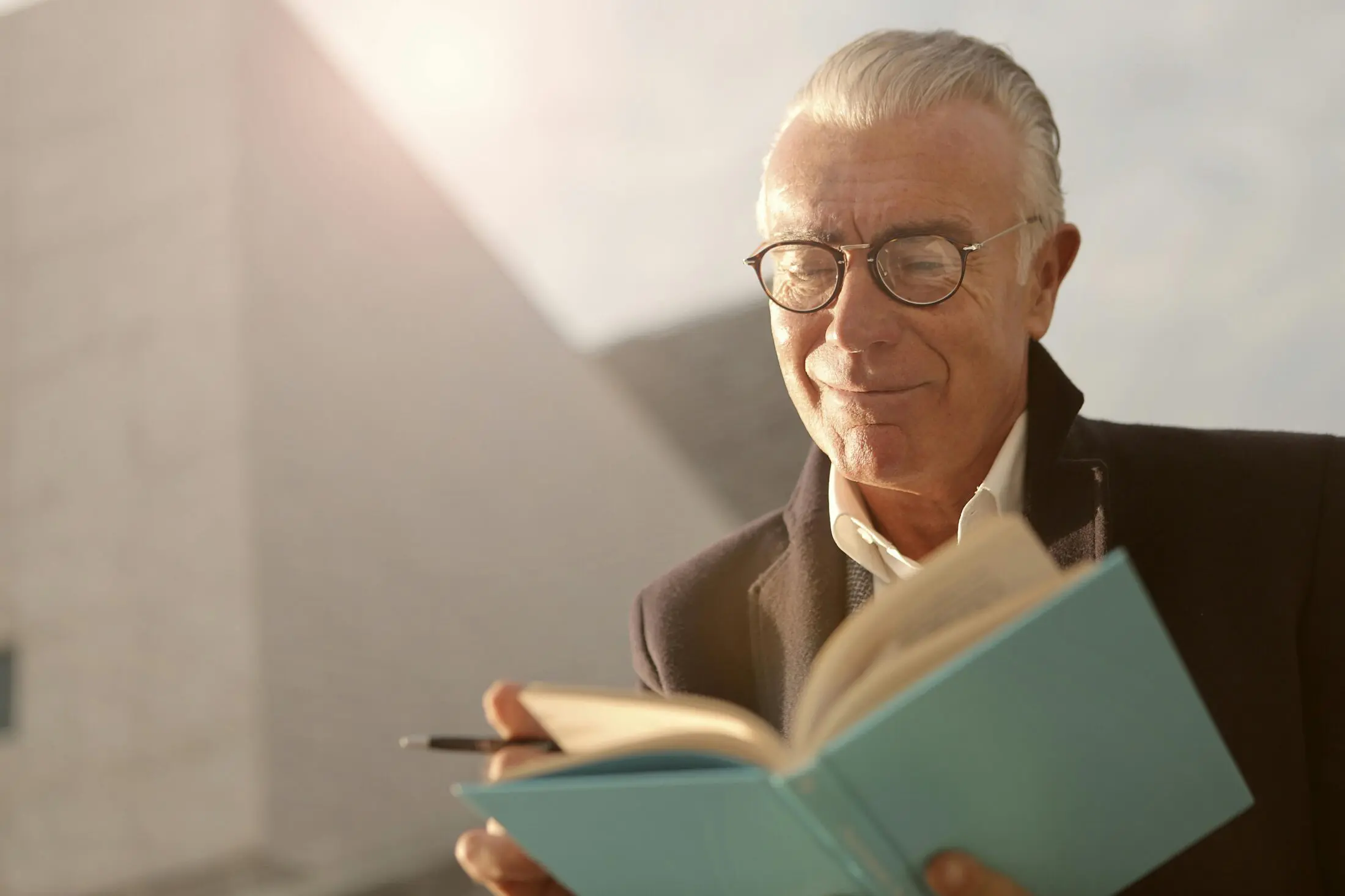 A handsome senior man with glasses, smiling and reading a blue book outdoors in the sunlight.