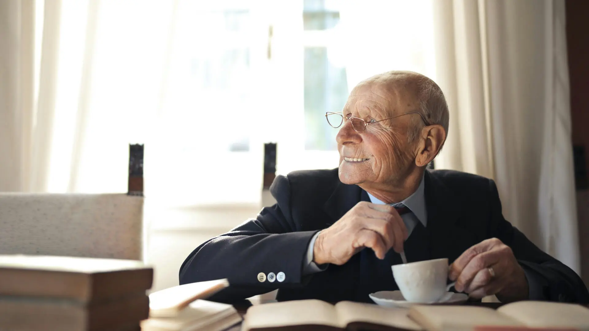A smiling senior man in a suit jacket stirring a cup of coffee at a table surrounded by open books.