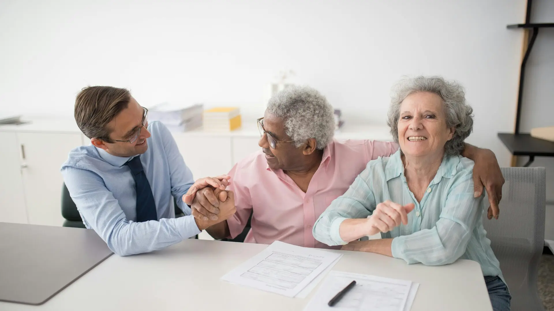 A professional financial advisor in a blue shirt and tie shakes hands with a smiling senior man of color. Beside him, an older woman with grey hair smiles warmly at the camera. They are sitting at a white desk with financial documents and a pen, appearing relieved and confident.