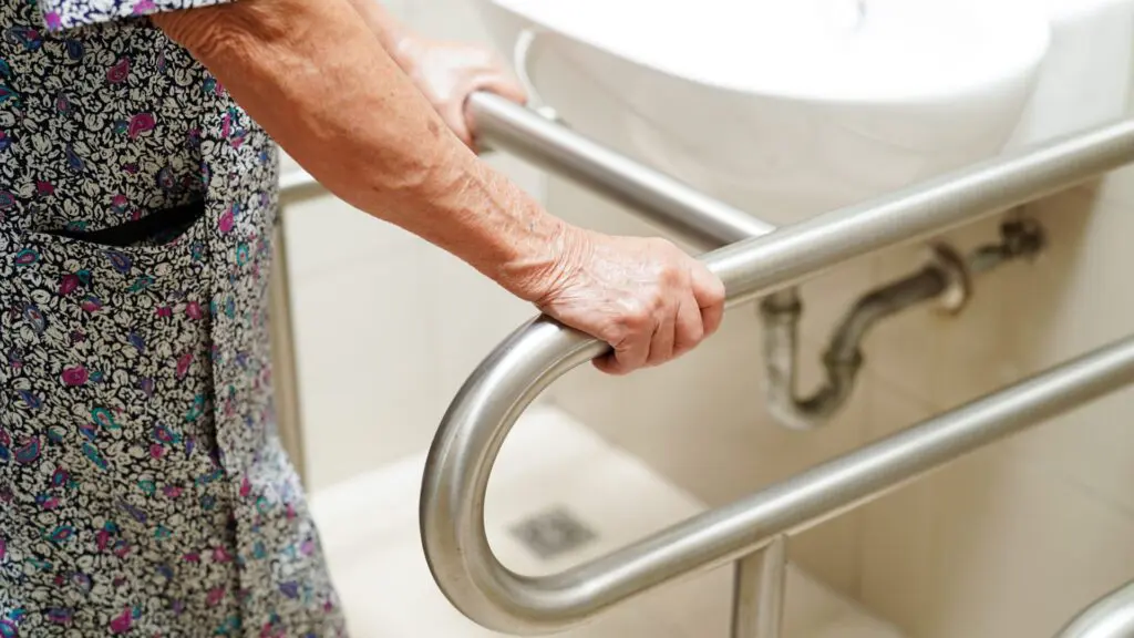 A close-up of a senior person's hands firmly gripping a sturdy metal grab bar installed in a bright bathroom next to a sink.