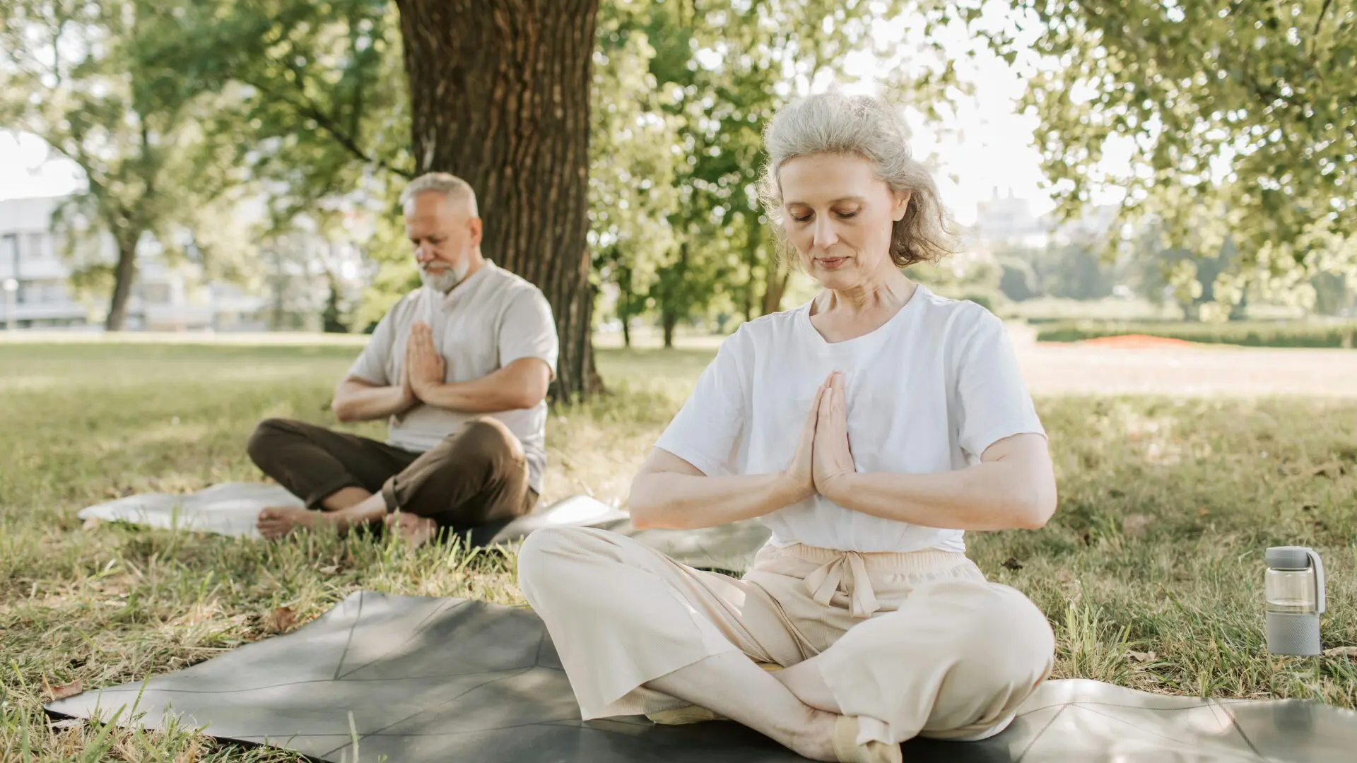 An older man and woman sitting in cross-legged meditation poses on yoga mats in a sunlit park, with hands in a prayer position and eyes closed for a peaceful mindfulness session.