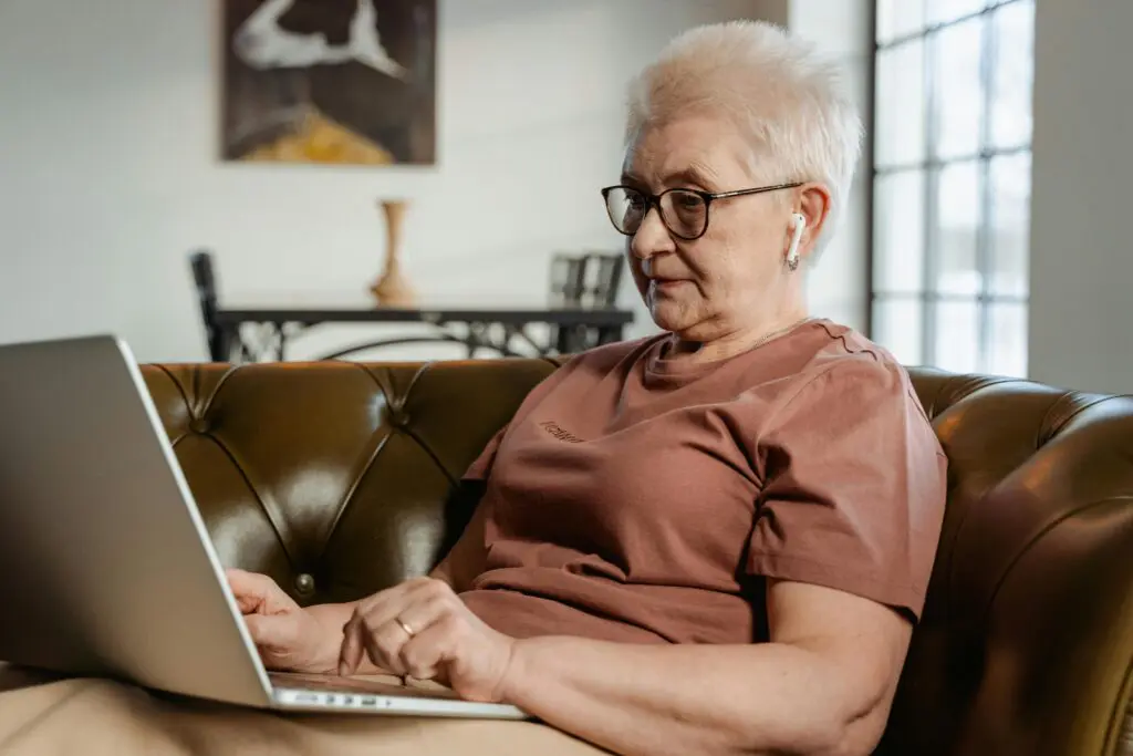 An older woman with short white hair and glasses sitting on a green leather sofa, focused on her laptop while wearing a wireless earbud. She is illustrating how digital hobbies like blogging or online learning enrich life after 60.
