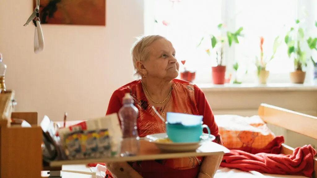 An elderly woman in a red shirt sitting comfortably in a well-lit room with a meal tray, representing a patient receiving personalized primary care.