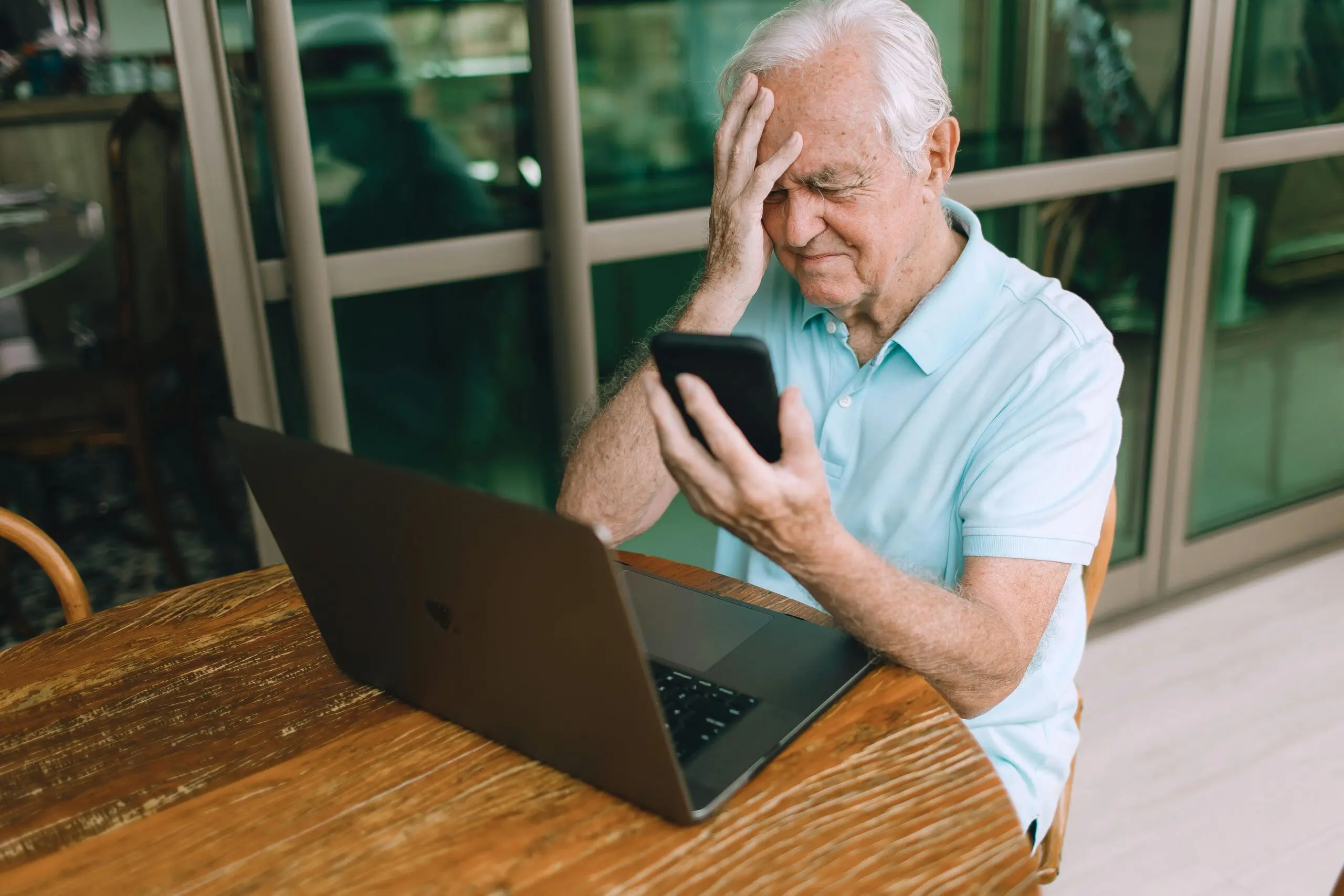 A stressed elderly man with a hand to his head, looking at his smartphone while sitting in front of an open laptop at a wooden table. The image is used to represent online challenges or scams for seniors.