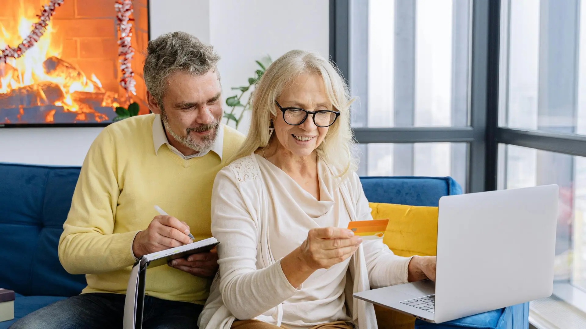 A senior couple sitting on a blue sofa, looking at a laptop together while the man takes notes in a binder and the woman holds a credit card. A warm fireplace is visible in the background.