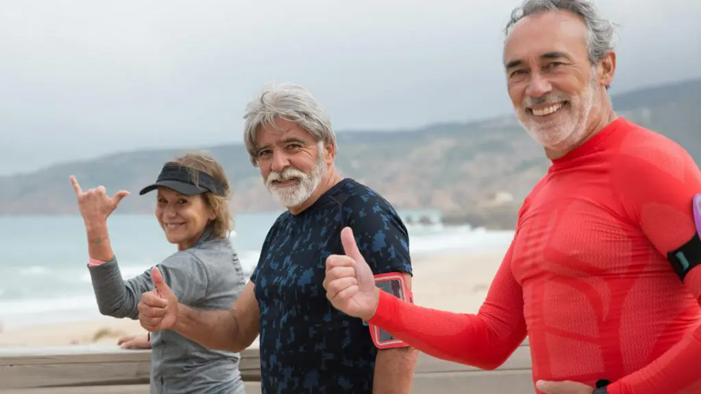 Three active seniors smiling and giving thumbs up at the beach, showcasing a positive growth mindset and healthy lifestyle.