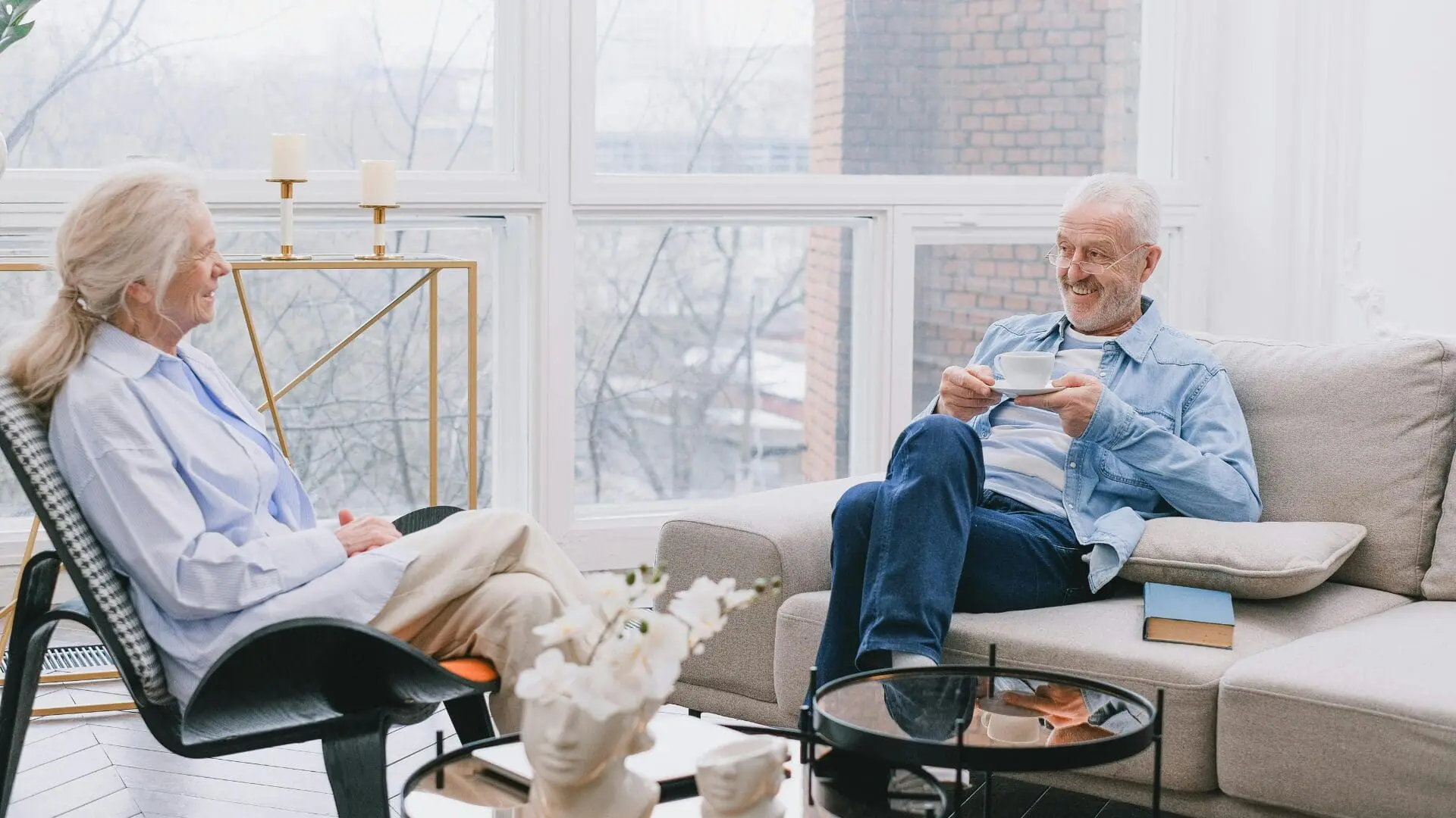 An elderly couple smiling and talking in a bright, modern living room with large windows, illustrating a healthy home environment for longevity.