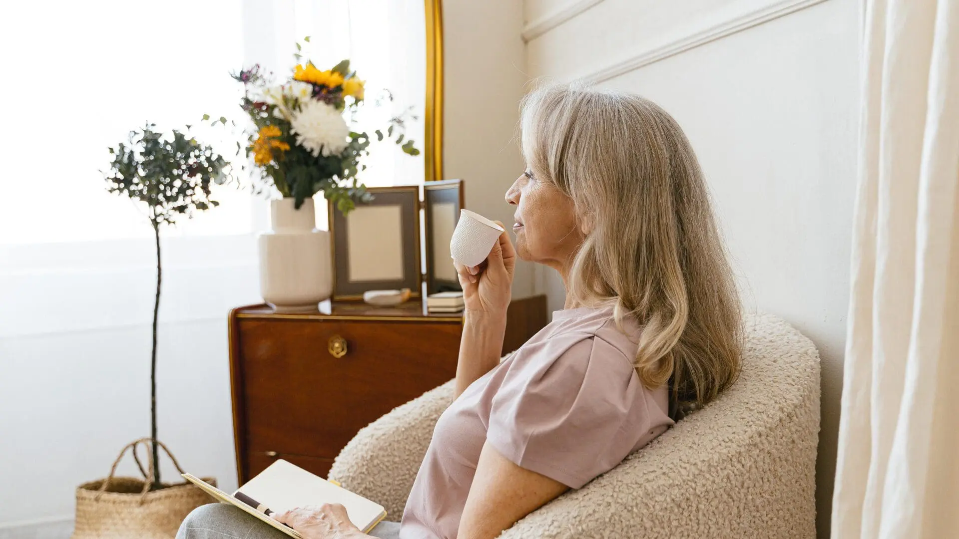Older woman with gray hair sits in a fluffy beige chair, holding a cup and writing in a notebook by a flowering vase and dresser in a bright room.