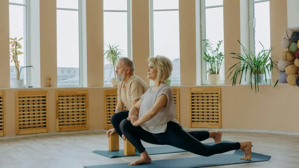 An older man and woman practicing a low lunge yoga pose on mats in a bright, sunlit studio with large windows and indoor plants.