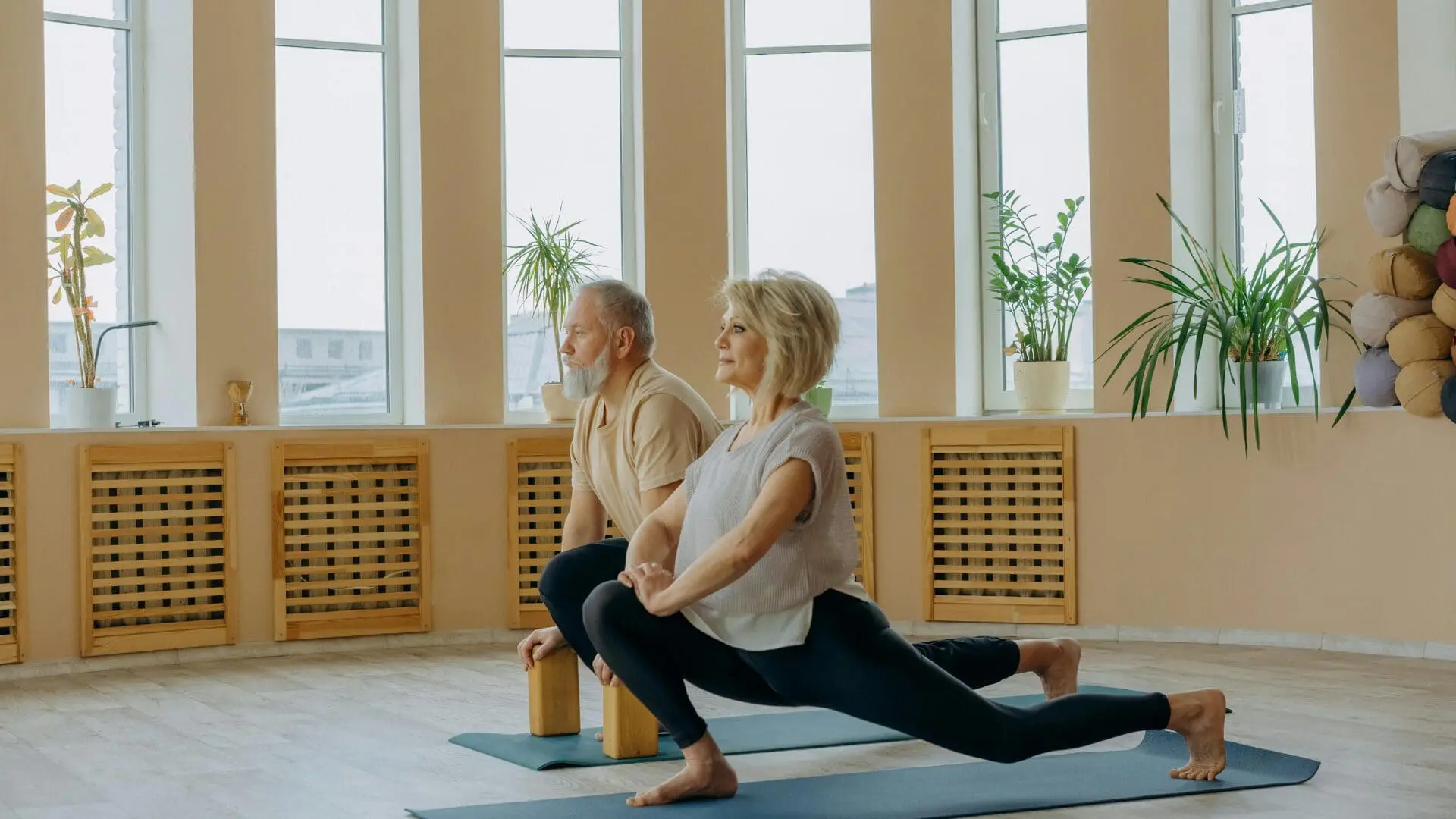 An older man and woman practicing a low lunge yoga pose on mats in a bright, sunlit studio with large windows and indoor plants.