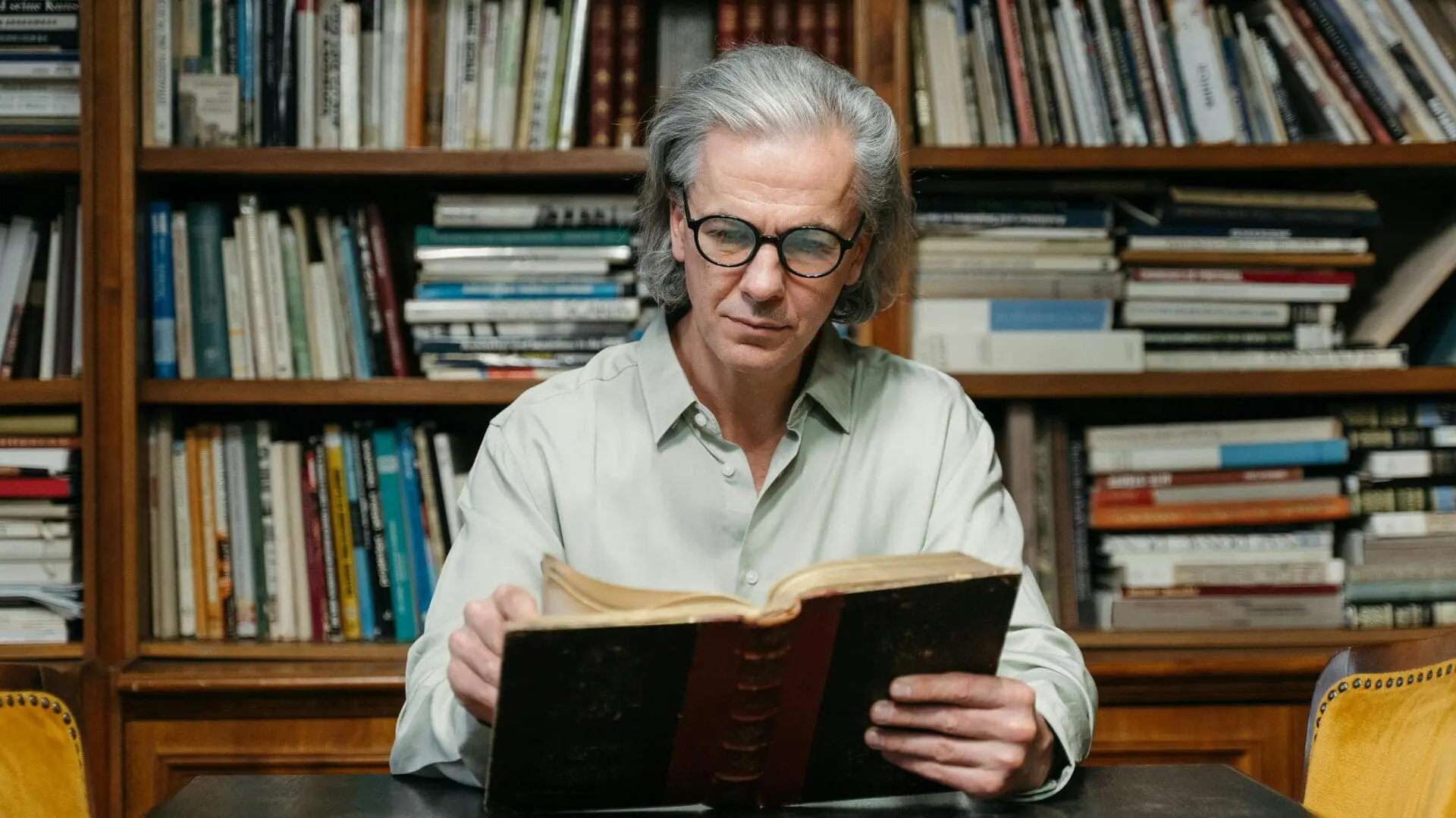 An older man with gray hair and glasses sitting at a table in a library reading a large hardcover book, with bookshelves in the background.