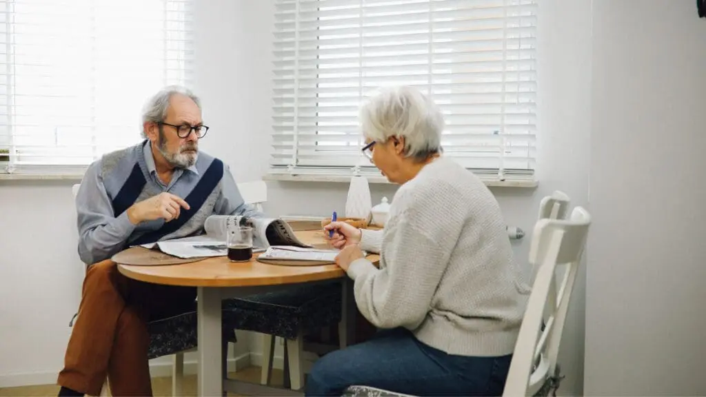 An elderly couple sitting at a wooden dining table by a window, reviewing a newspaper and paperwork together in a bright, modern room.
