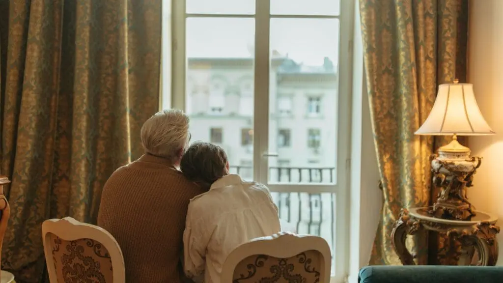 An older couple, viewed from behind, sits close together looking out a large window of their comfortably furnished home at a city view.