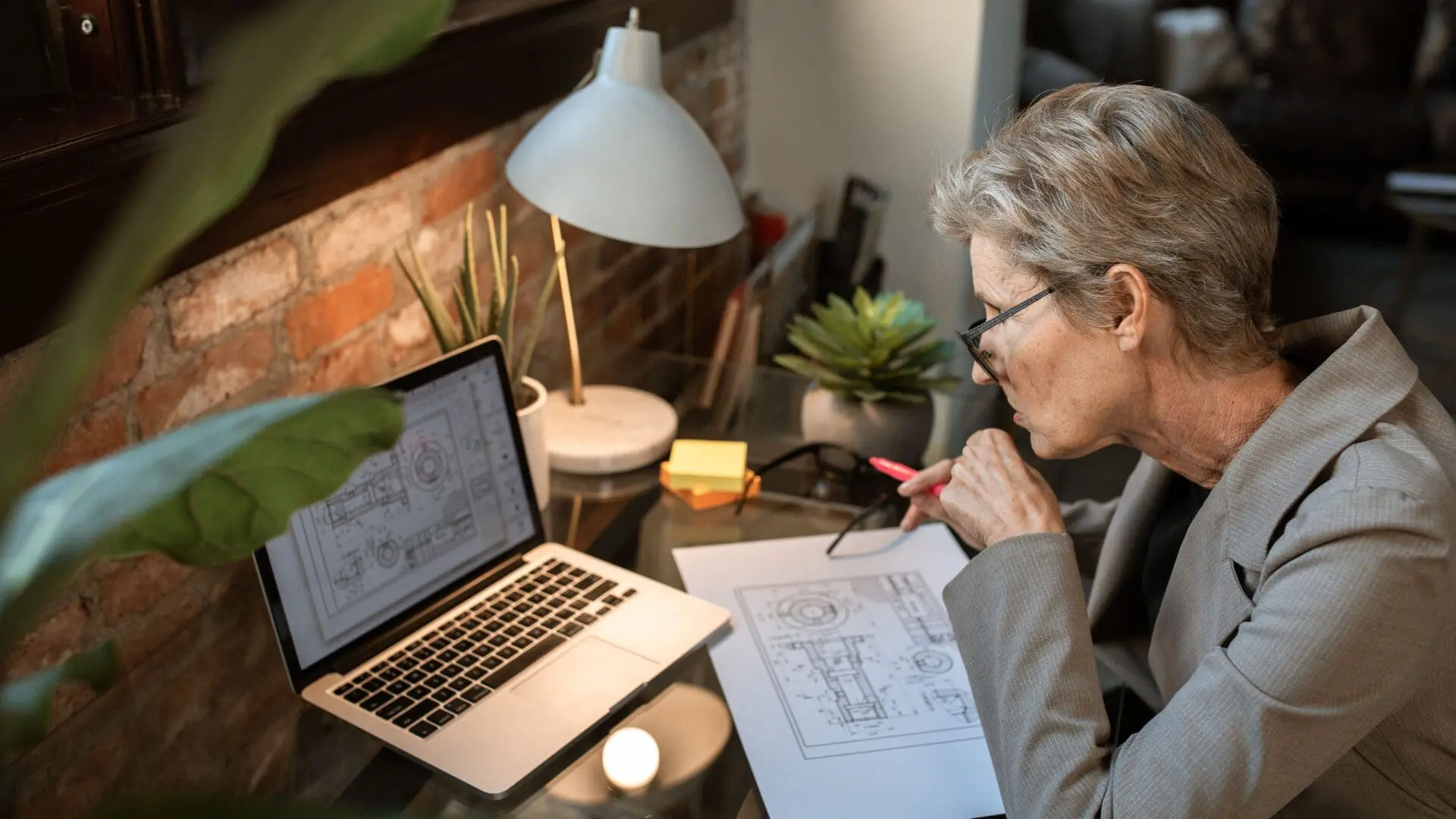 An older woman with short grey hair and glasses working at a desk, reviewing a technical blueprint drawing on paper and comparing it to a design on her laptop screen.