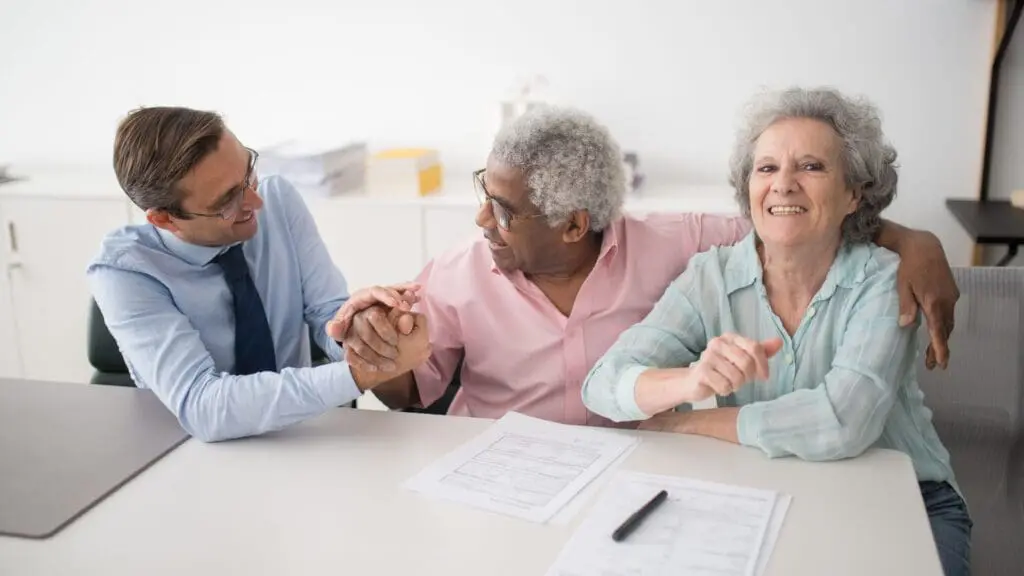 An insurance agent shaking hands with an elderly man sitting next to his smiling wife at a desk during a meeting about insurance options for older adults.