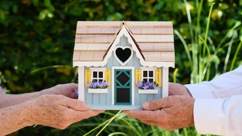 Two pairs of aging hands gently holding a small, decorative model of a blue house with a wooden roof, a heart-shaped window, and purple flowers in the window boxes against a blurred green background.