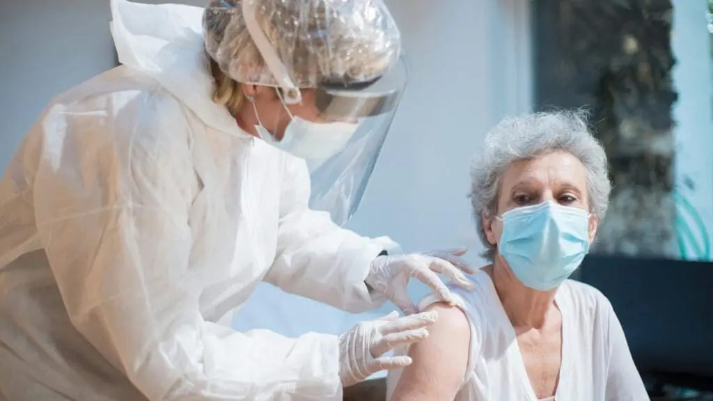 Healthcare worker in full PPE and face shield administers a vaccine to an elderly woman wearing a mask or shielding her arm for the injection.