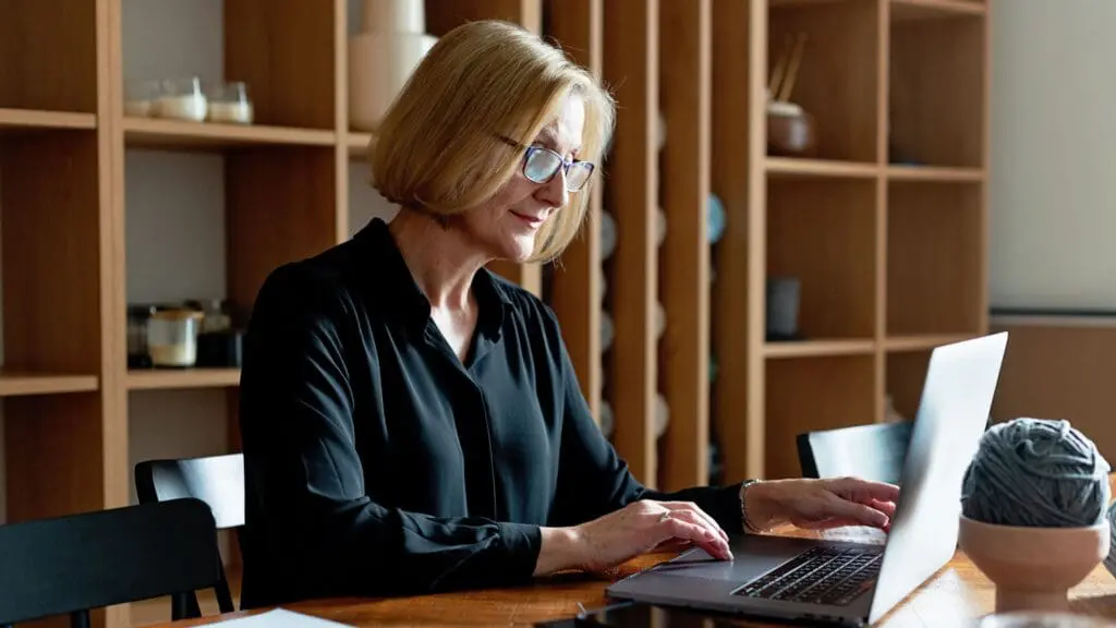 An older adult woman with blonde hair and glasses smiling while working on a laptop at a wooden table at home.