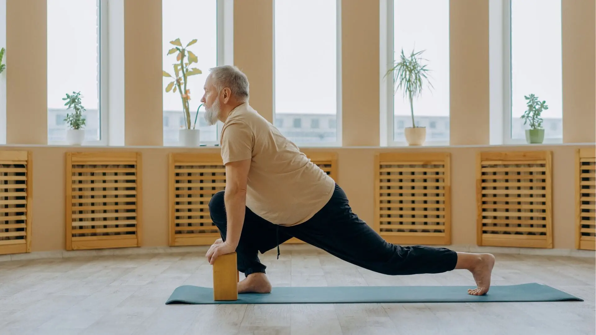 A senior man with a gray beard practicing a low lunge stretch on a blue yoga mat in a bright, spacious room. He is utilizing a wooden yoga block for stability and support, focusing on flexibility.