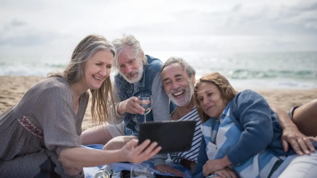 A group of four smiling, retired friends gathering together on a sandy beach blanket, laughing while looking at a digital tablet.