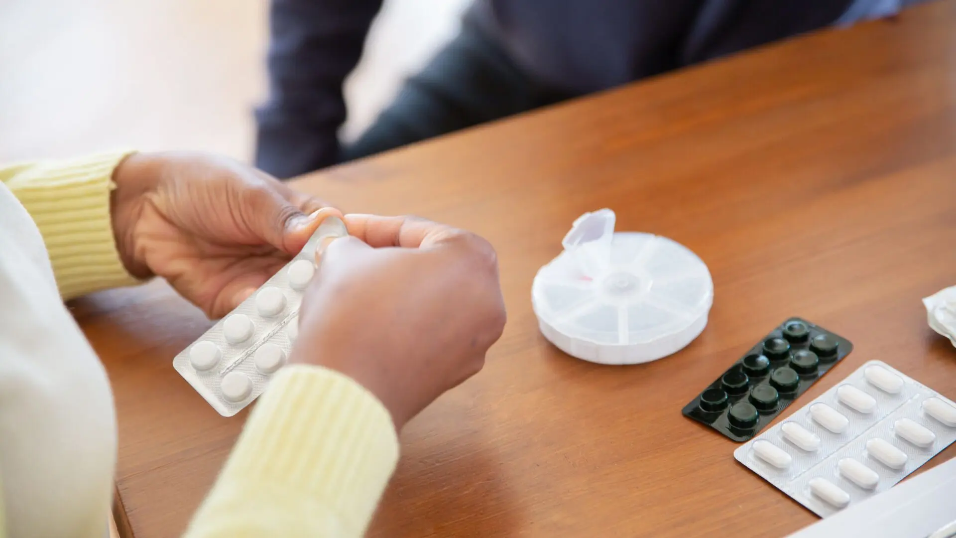 Older adult’s hands pressing a pill out of a blister pack at a wooden table, next to a weekly pill organizer and more blister packs of medication.