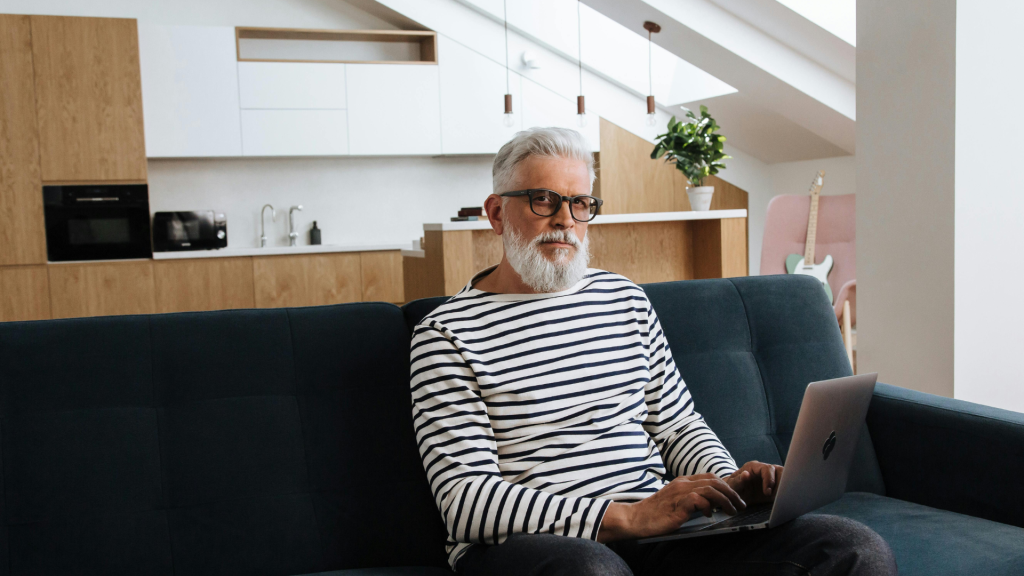 A senior man with a white beard and glasses using a laptop while sitting on a couch.