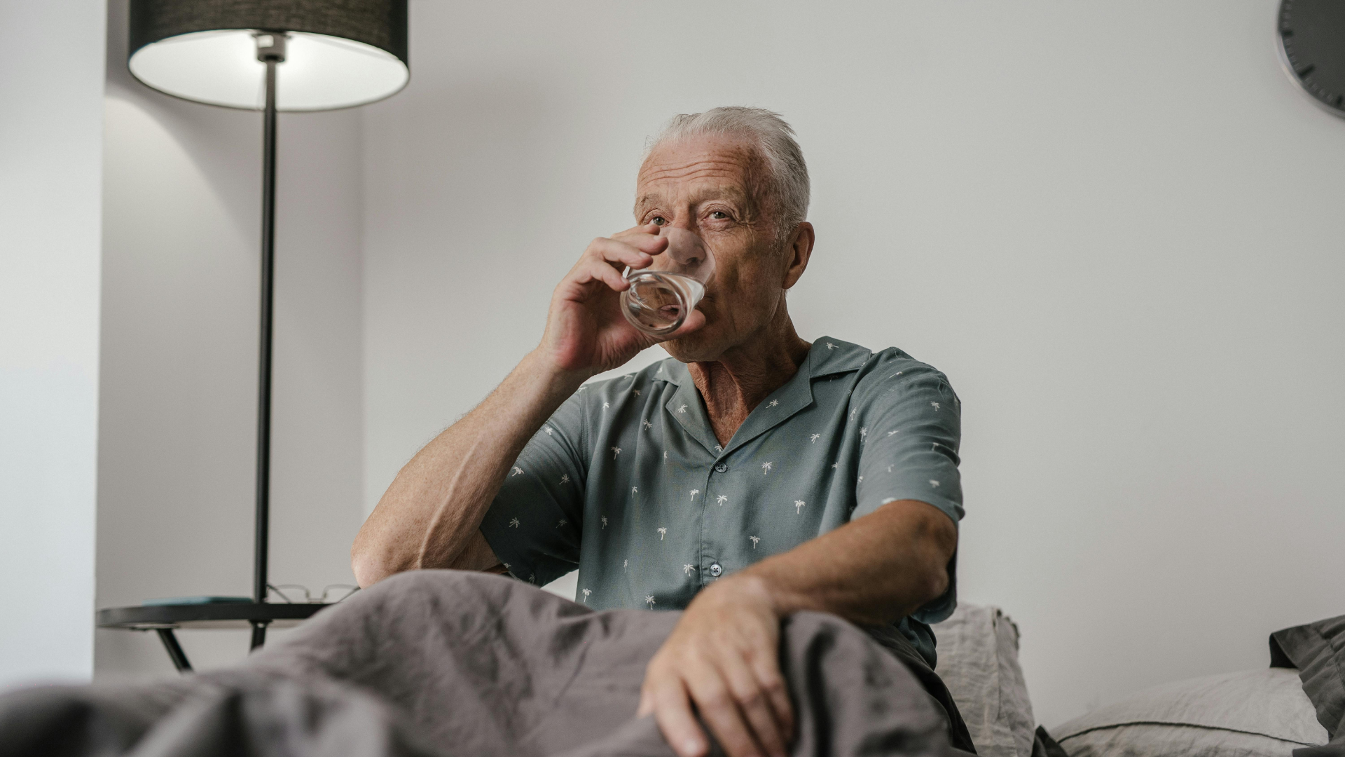 A senior man in a gray shirt drinking a glass of water while sitting up in bed.