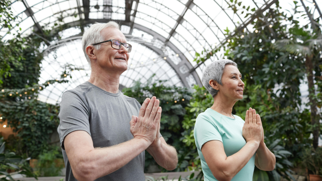 A senior man and woman with hands in prayer position, practicing yoga or meditation in a lush indoor garden setting.