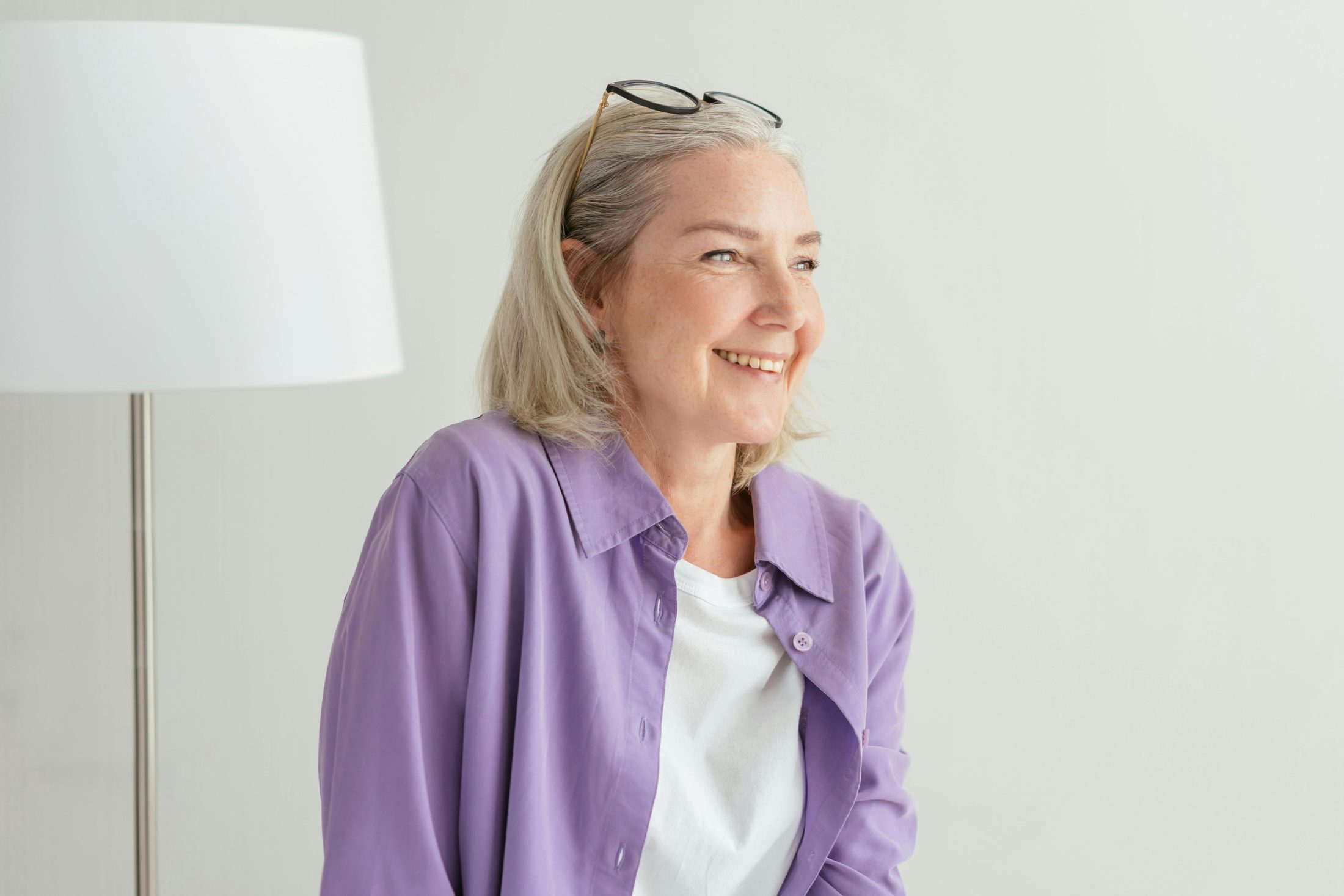 A smiling senior woman with blonde hair, wearing a purple shirt and glasses on her head, looking happily off to the side.