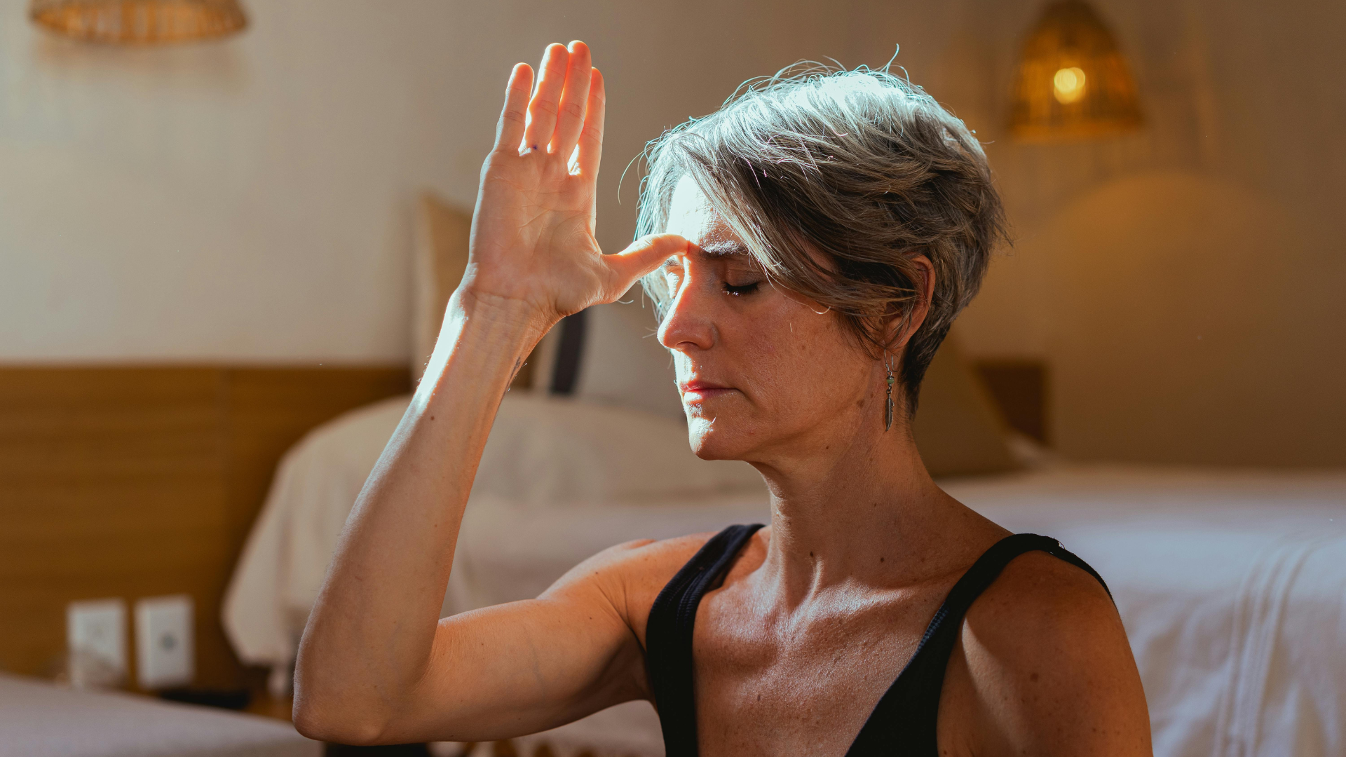 A senior woman with short gray hair practicing alternate nostril breathing (Nadi Shodhana Pranayama) indoors.