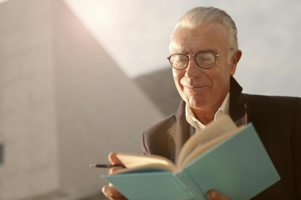 A handsome senior man with glasses, smiling and reading a blue book outdoors in the sunlight.