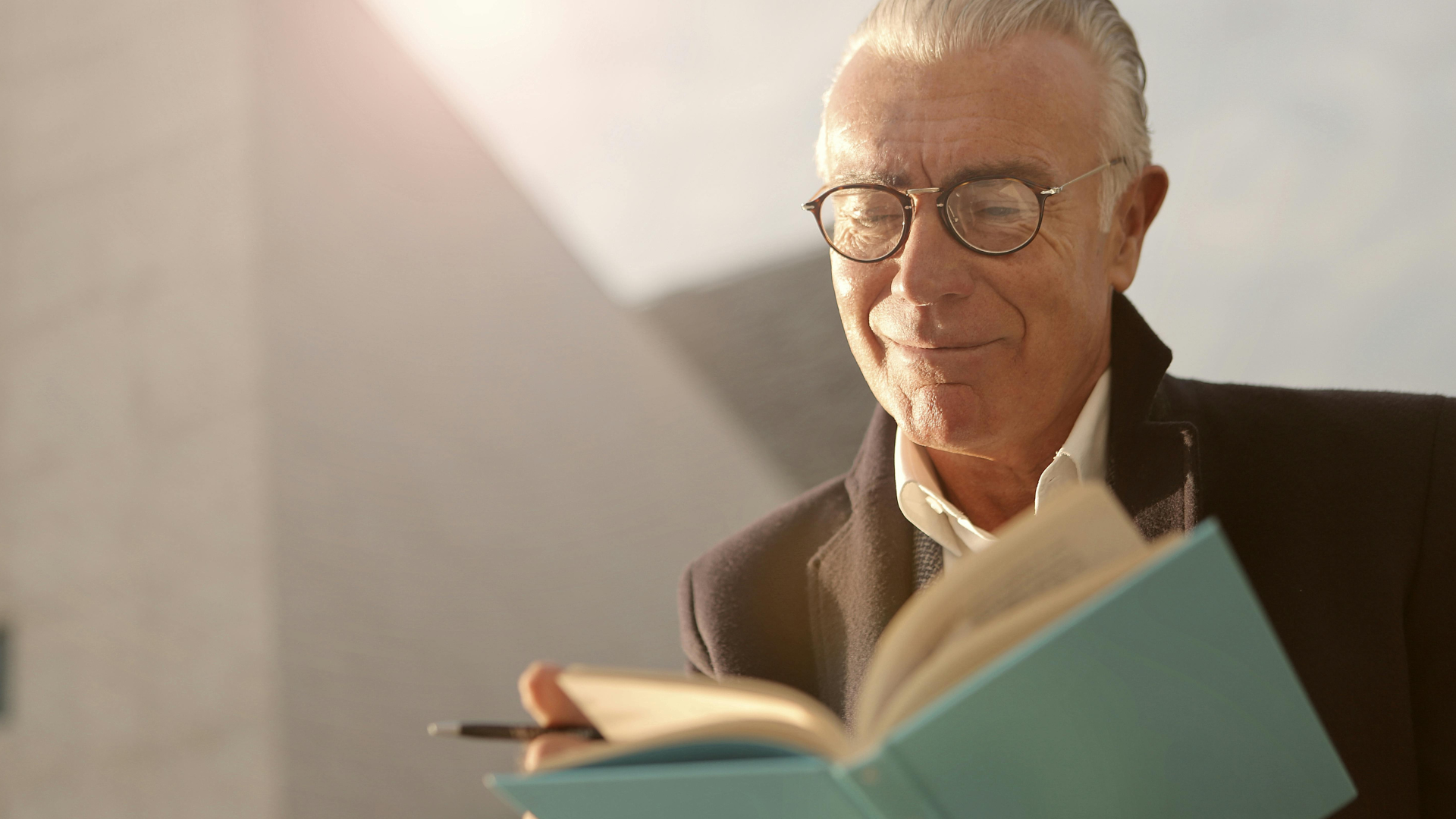 A handsome senior man with glasses, smiling and reading a blue book outdoors in the sunlight.